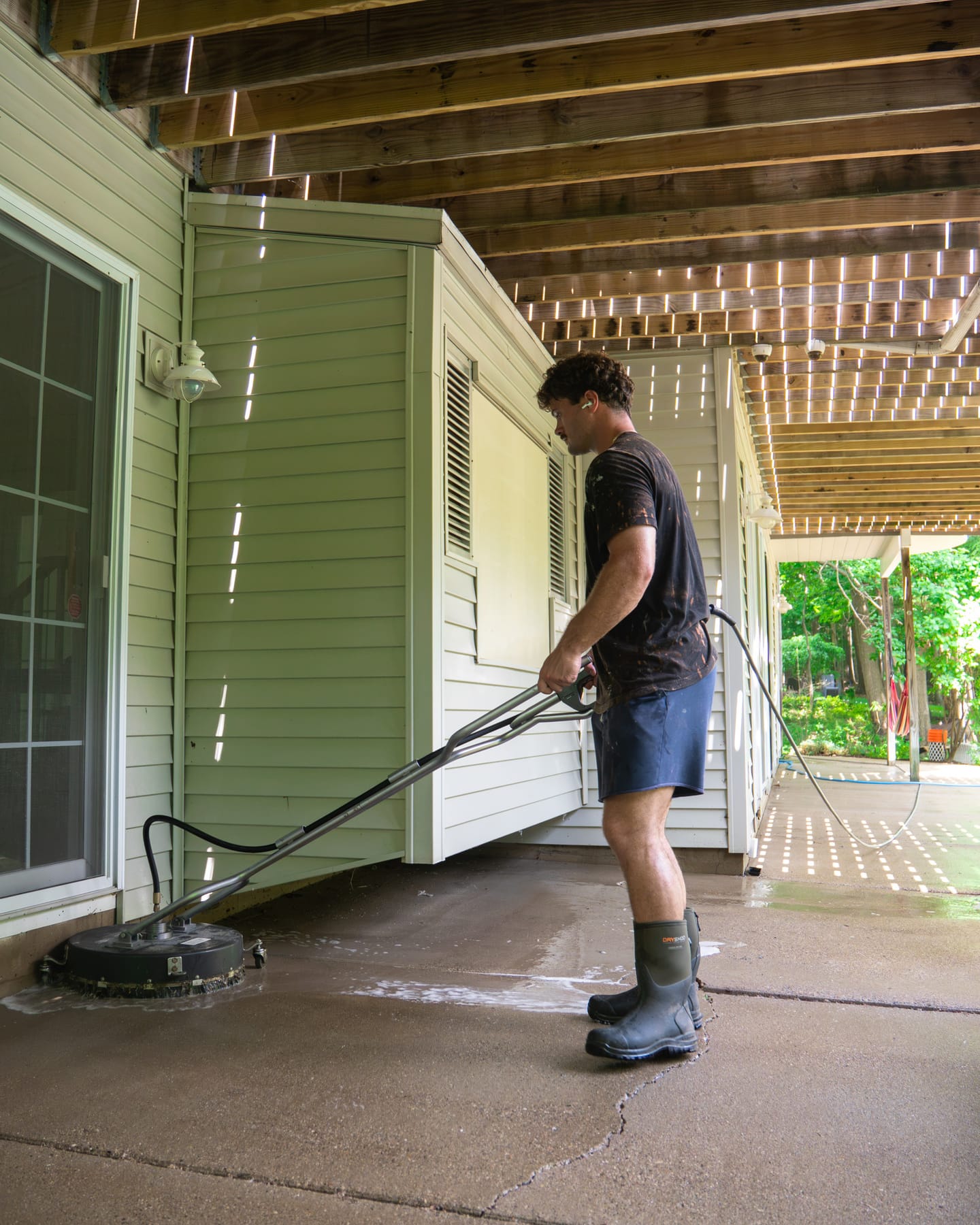 Technician running a rotary surface cleaner across a shaded concrete patio beneath a raised deck