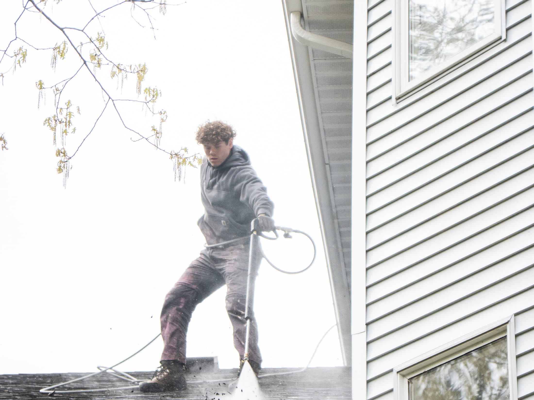 Technician clearing a home's gutters from the roof, debris flushing out over the roof edge