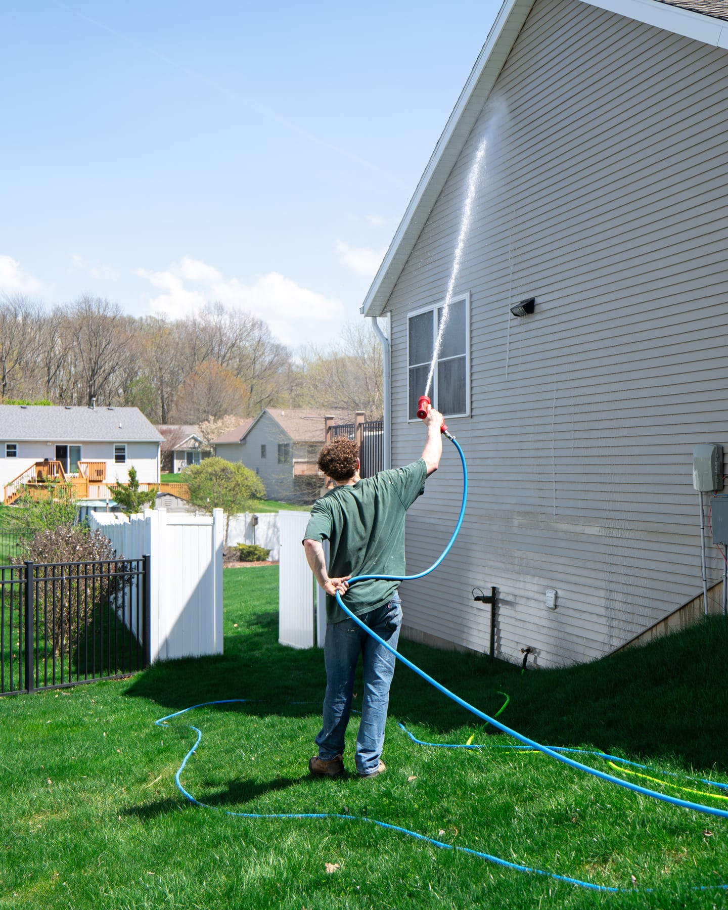 Technician soft-washing the gable end of a two-story home from ground level, no ladder against the house