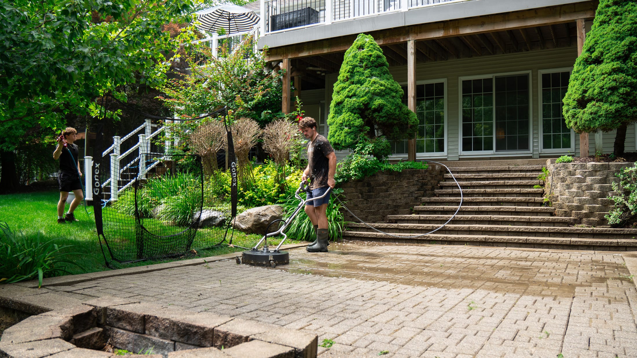 Technician running a rotary surface cleaner across a large brick paver patio behind a two-story home