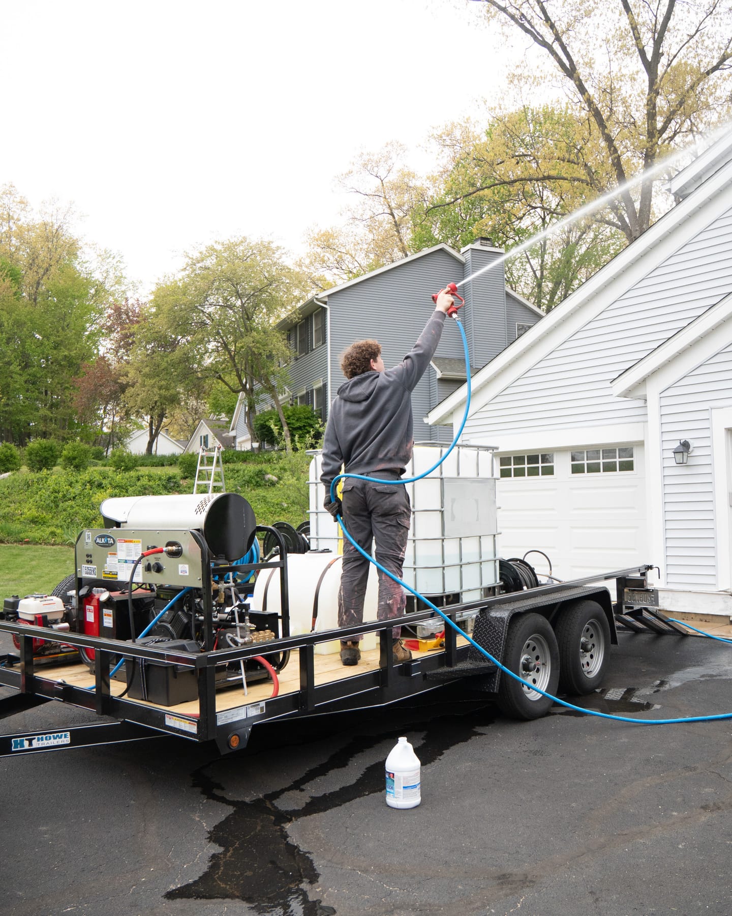 Technician soft-washing a roofline from the trailer rig, with pressure washer, water tank and hose reel aboard